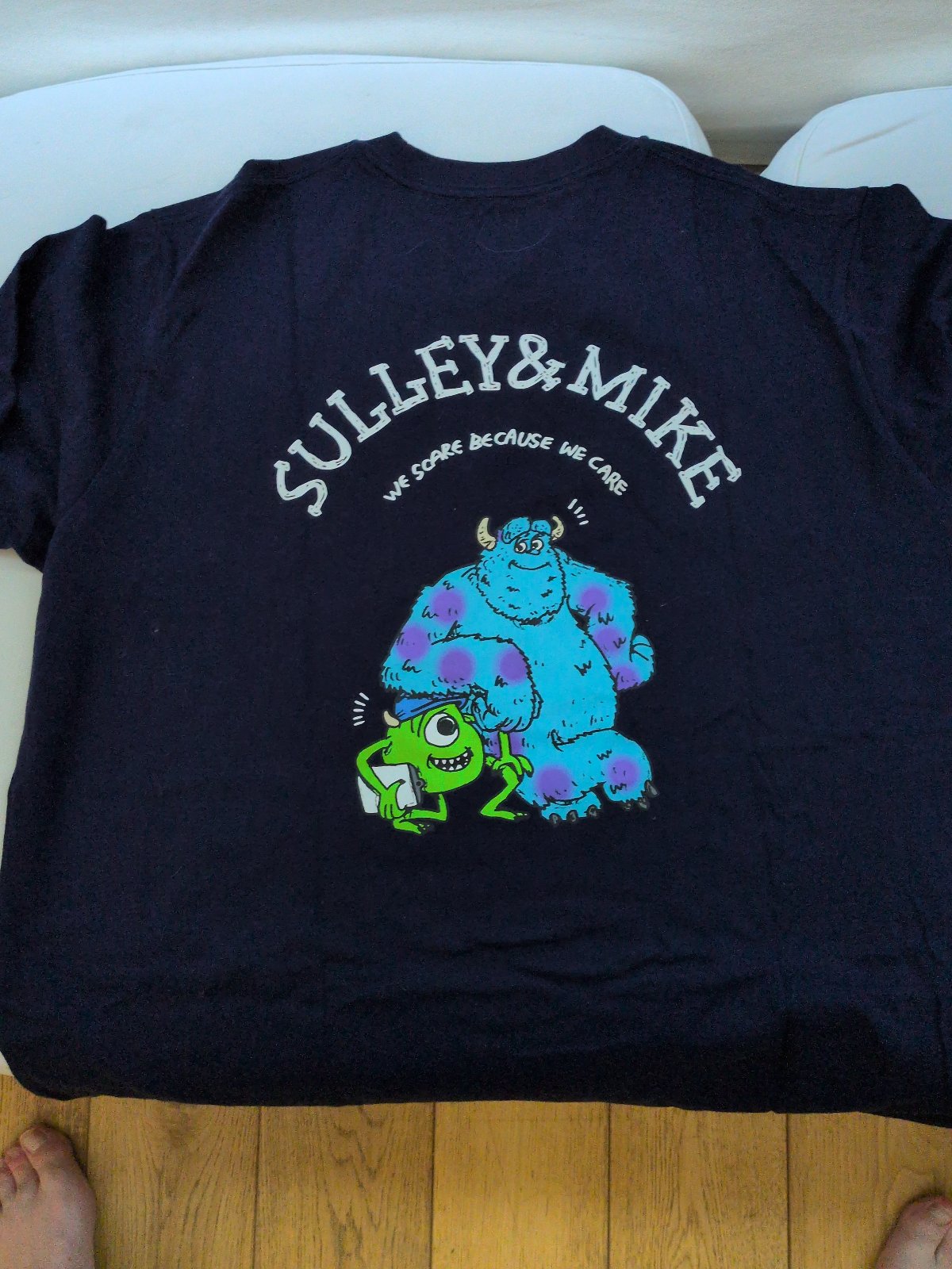 Navy blue short-sleeved t-shirt with a small printed monster-factory door on the chest and, on the reverse, the arched text "SULLEY&MIKE / WE SCARE BECAUSE WE CARE" above cartoon blue and green monsters.
